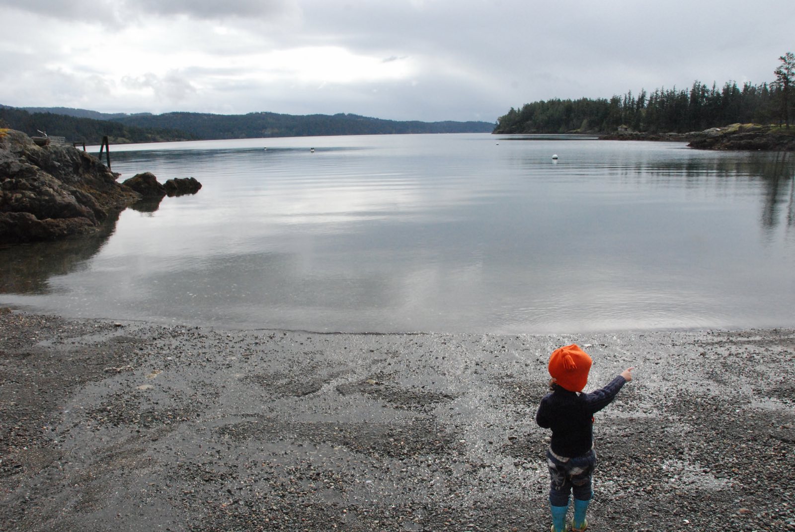 A small child in an orange hat stands on a pebble beach, pointing out across a still, grey cove.